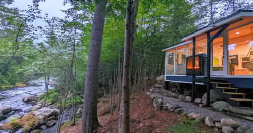 A modern cabin by a river, surrounded by trees, with large windows reflecting the natural scenery.