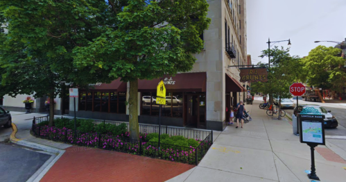 Exterior view of a restaurant with outdoor seating, surrounded by trees and flowers, on a city street corner.