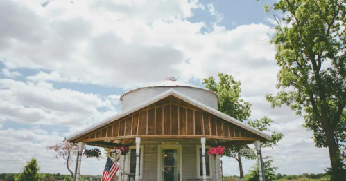 A charming round house with a porch, surrounded by greenery and a blue sky dotted with clouds. An American flag is visible.