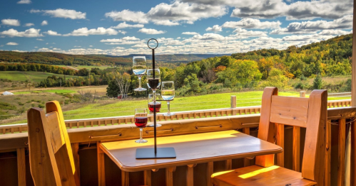 A scenic view of rolling hills and colorful trees from a wooden deck with a table and wine glasses.