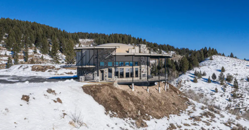 Modern house on a snowy hillside, surrounded by trees and mountains under a clear blue sky.