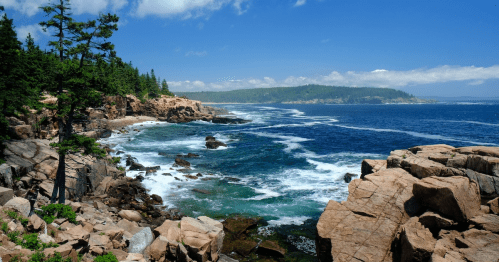 A scenic coastal view with rocky shores, lush greenery, and waves crashing against the rocks under a clear blue sky.