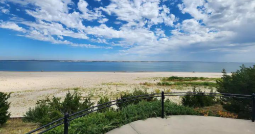 A scenic view of a beach with a calm sea, blue sky, and scattered clouds, framed by greenery and a railing.