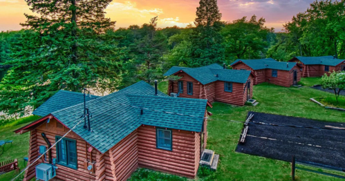 Aerial view of rustic log cabins surrounded by greenery at sunset, with a river in the background.