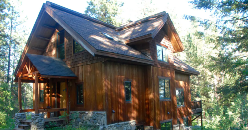 A wooden house with a sloped roof, surrounded by trees and sunlight filtering through the branches.