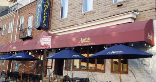 Exterior of a casual eatery with a maroon awning, outdoor seating, and blue umbrellas, featuring a stone and brick facade.