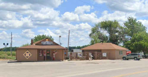A small brick building with a sign, surrounded by trees and a clear blue sky, located on a rural road.