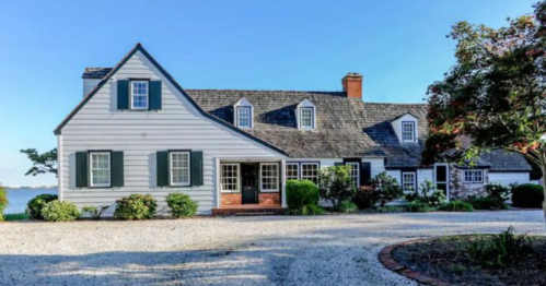 A charming gray house with a sloped roof, surrounded by greenery and a gravel driveway, near a body of water.
