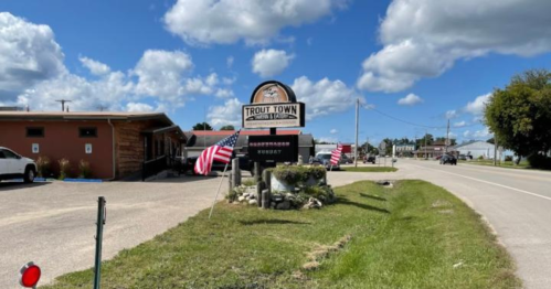 Sign for Trout Town with American flags, surrounded by greenery, on a sunny day with clouds in the sky.