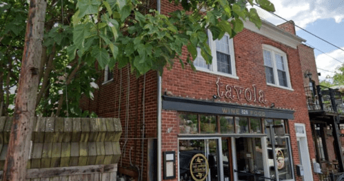 A brick building with a sign reading "Tavola" and large windows, surrounded by trees and a wooden fence.