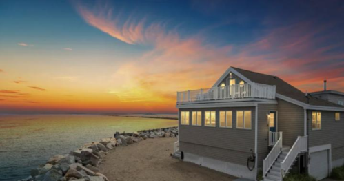 A coastal house at sunset, with a rocky shoreline and vibrant sky colors reflecting on the water.