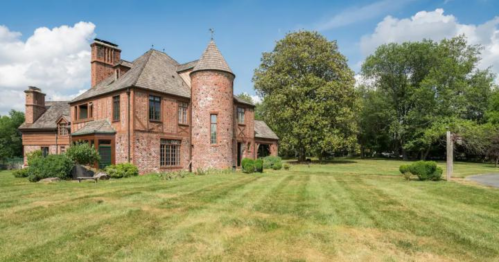 A large, unique brick house with a turret, surrounded by a green lawn and trees under a blue sky with clouds.