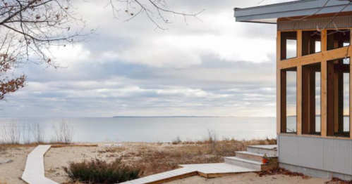 A serene beach scene with a modern house, sandy path, and calm water under a cloudy sky.