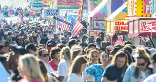 A crowded street fair with people walking among colorful booths and American flags, enjoying food and drinks.