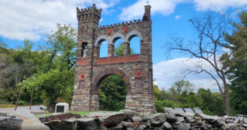 Stone archway with towers, surrounded by trees and blue sky, showcasing historical architecture in a natural setting.
