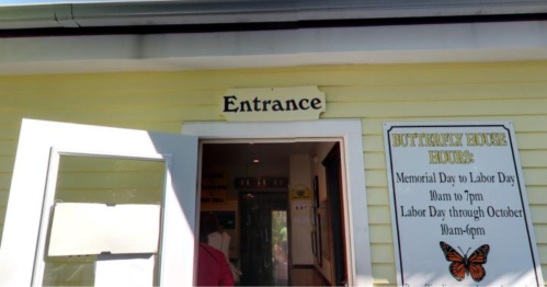A yellow building with an "Entrance" sign and a nearby hours sign for a butterfly house.