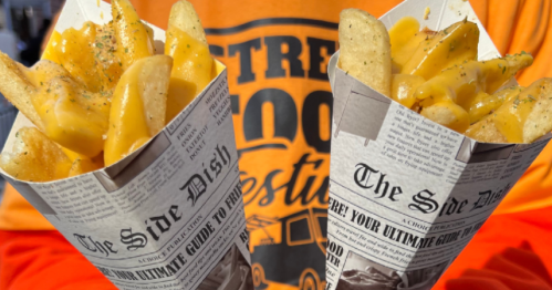 Two cones of fries topped with cheese sauce, held by a person in an orange shirt with "Street Food Festival" printed on it.