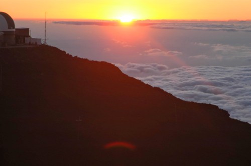 A stunning sunset over a mountain peak, with clouds below and a silhouette of an observatory on the left.