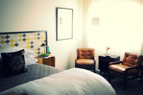 Cozy bedroom with a bed, two brown chairs, a nightstand, and a window letting in natural light.