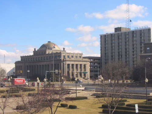 Historic building with a dome, surrounded by modern structures and a clear blue sky. Green landscaping in the foreground.