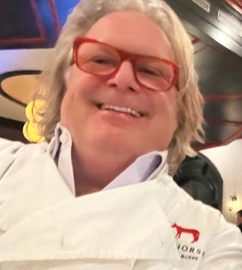 A smiling person with curly hair and red glasses, wearing a white chef's coat, poses for a selfie in a restaurant.