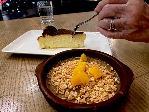 A hand holding a fork above a slice of cheesecake on a plate, with a dessert of chocolate mousse and orange segments in the foreground.