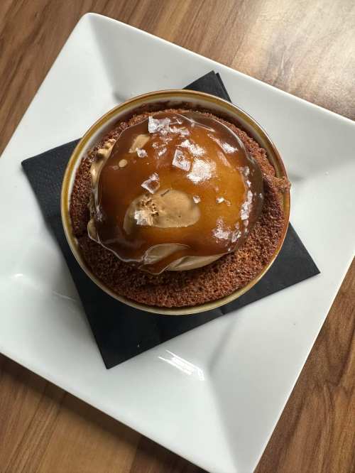 A round dessert with a glossy caramel topping, sprinkled with sea salt, on a black napkin and white plate.