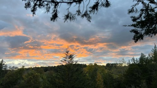 A vibrant sunset with orange hues peeking through clouds over a forested landscape.