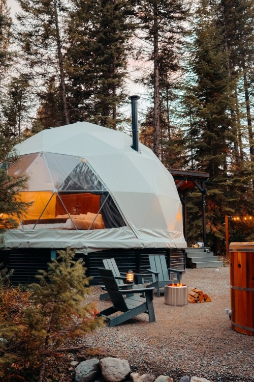 A cozy geodesic dome cabin surrounded by trees, featuring a wood stove and outdoor seating with lanterns.
