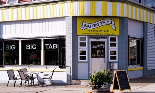 Exterior of Big Big Table Community Cafe, featuring large windows, a bright yellow sign, and outdoor seating.