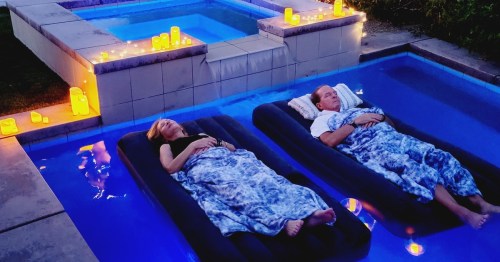 A couple relaxes on floating loungers in a pool, surrounded by glowing candles and a serene atmosphere at dusk.