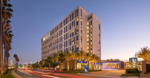 Modern hotel building at dusk, surrounded by palm trees and city lights, with traffic passing by.