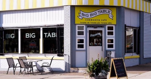 Exterior of "Big Big Table" community café with yellow awning, open sign, and outdoor seating.