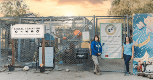 Two women stand by a fenced entrance of Animal Tracks Inc., with a colorful mural and banners in the background.
