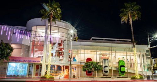 Exterior of the Muckenthaler Art Museum at night, featuring colorful cars displayed on the facade and palm trees.