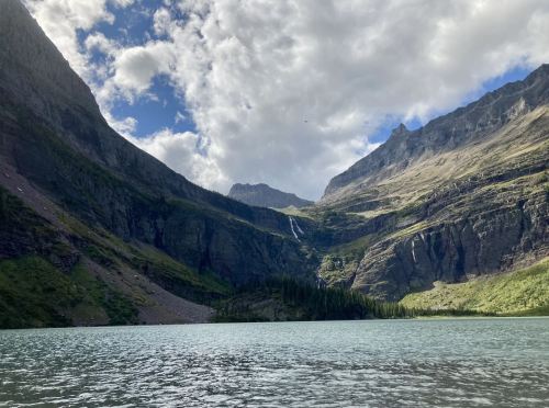 A serene lake surrounded by towering mountains and a waterfall cascading down the rocky cliffs under a cloudy sky.