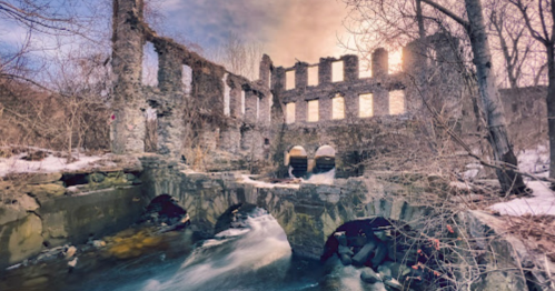 Ruins of a stone building beside a flowing stream, surrounded by bare trees and a winter landscape.