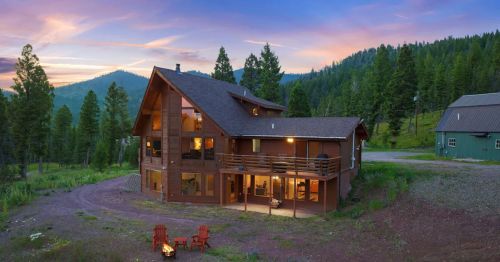 A large wooden house surrounded by trees, with a sunset sky and a fire pit in the foreground.