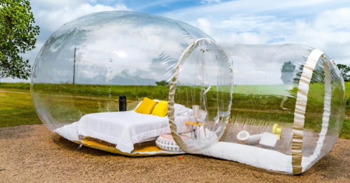 A transparent bubble structure with a bed and seating, set in a grassy area under a blue sky.