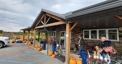 A rustic store entrance with pumpkins and decorations outside, featuring two people walking by on a cloudy day.