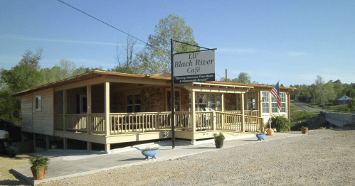 A wooden café building with a porch, sign reading "Lil' Black River Café," surrounded by greenery and gravel.