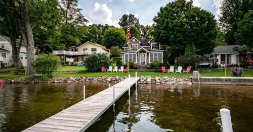 A serene lakeside view featuring a wooden dock, colorful chairs, and a charming house surrounded by trees.