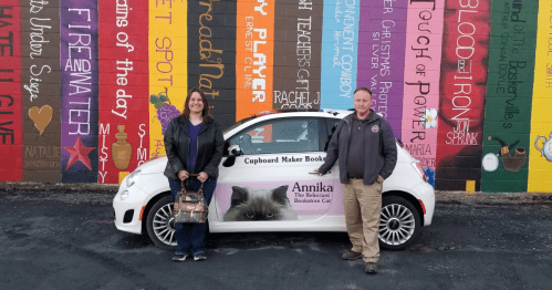 A woman and a man stand beside a car featuring a cat graphic, in front of a colorful mural.
