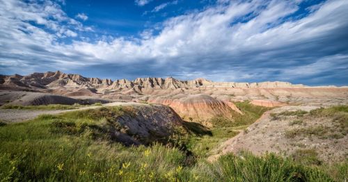 A scenic view of colorful rock formations under a blue sky with clouds, surrounded by green grass and shrubs.