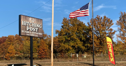Sign for "The Hitching Post RV Park & Tiny Homes" with an American flag and colorful "Now Open" banner in autumn scenery.