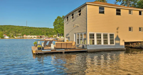 A lakeside house with large windows and a deck, surrounded by water and greenery under a clear blue sky.