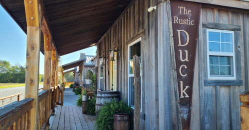 A wooden building with a sign reading "The Rustic Duck," featuring a porch and barrels, under a clear blue sky.