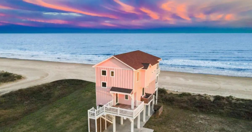 A pink beach house stands near the shore, with vibrant sunset colors in the sky and calm ocean waves in the background.