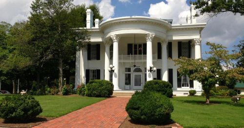 A grand white mansion with columns, surrounded by green lawns and trees under a partly cloudy sky.