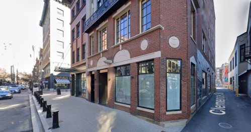 A brick building at a street corner with large windows and decorative elements, surrounded by a cityscape.